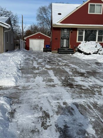A red house with a snow-covered driveway, a detached garage, and patches of ice and snow on a bright, sunny winter day.