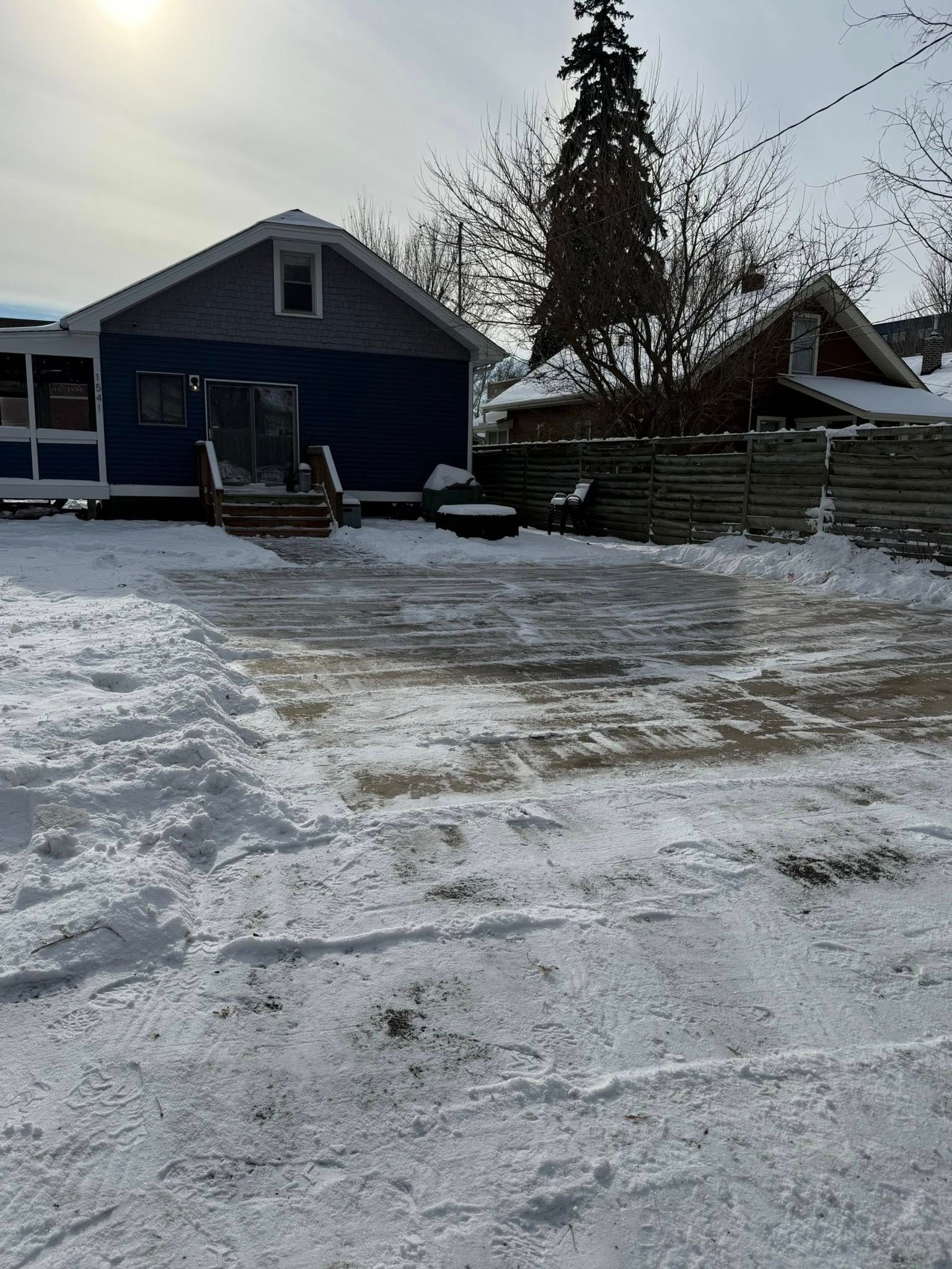 Snow-covered backyard with a blue house, a wooden deck, and a neighboring home under a bright sun.