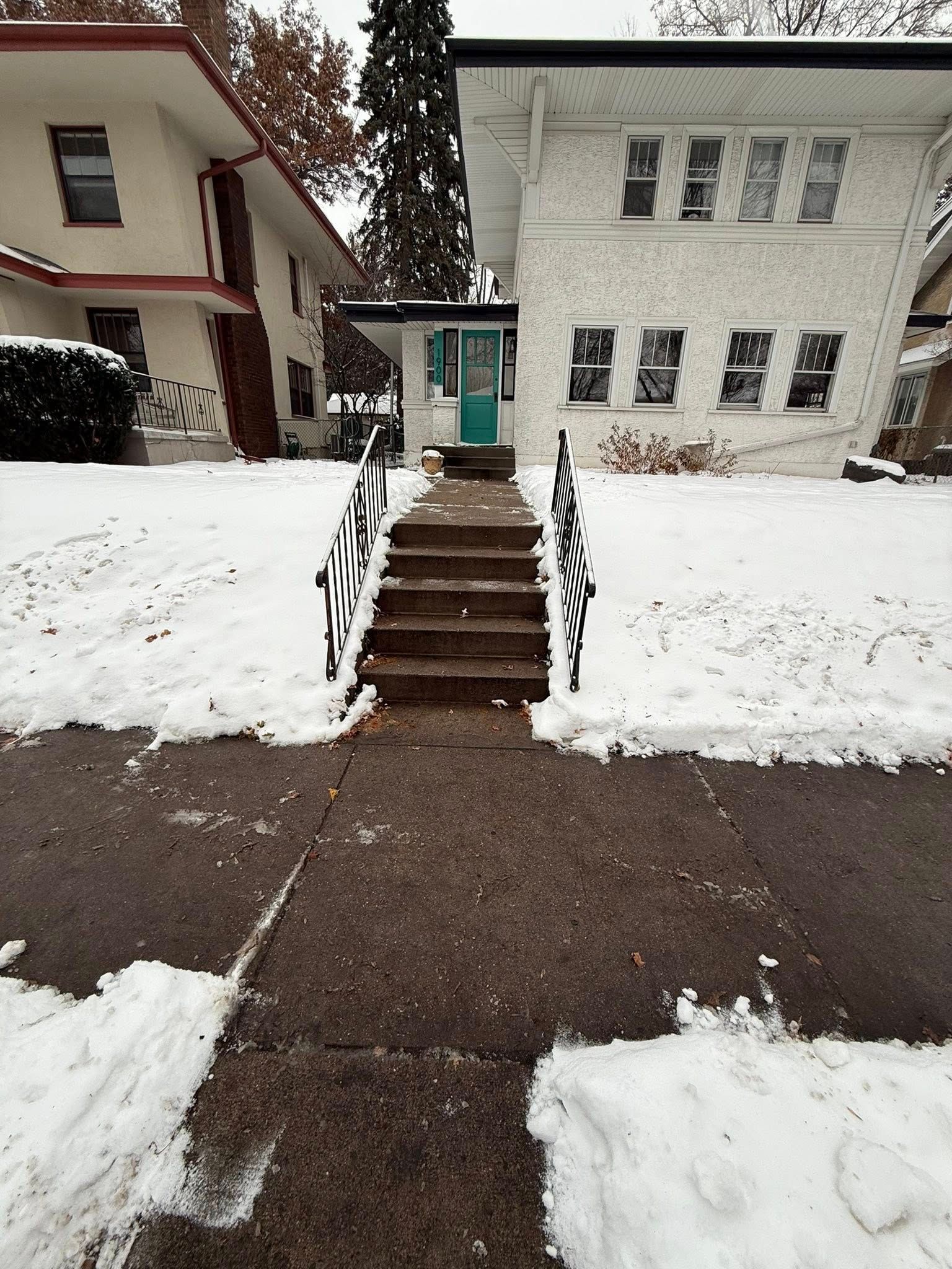 A snow-covered front yard and concrete walkway leading to a set of stairs and a teal front door of a two-story home.