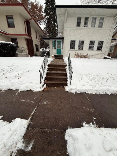 A snow-covered front yard and concrete walkway leading to a set of stairs and a teal front door of a two-story home.