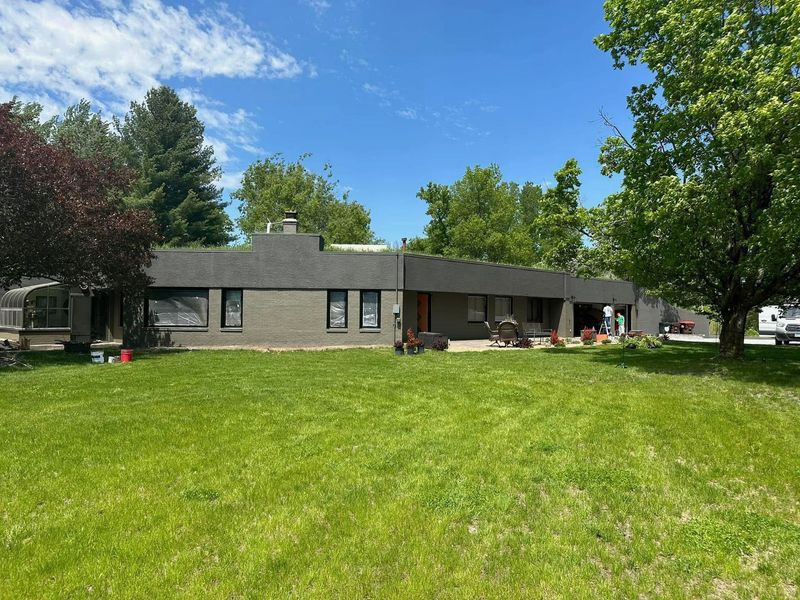 A single-story, dark gray brick ranch house with a flat roof, surrounded by a large green lawn under a bright blue sky.