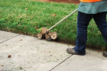 A worker in an orange vest uses a wheeled edger to trim grass along a concrete sidewalk.