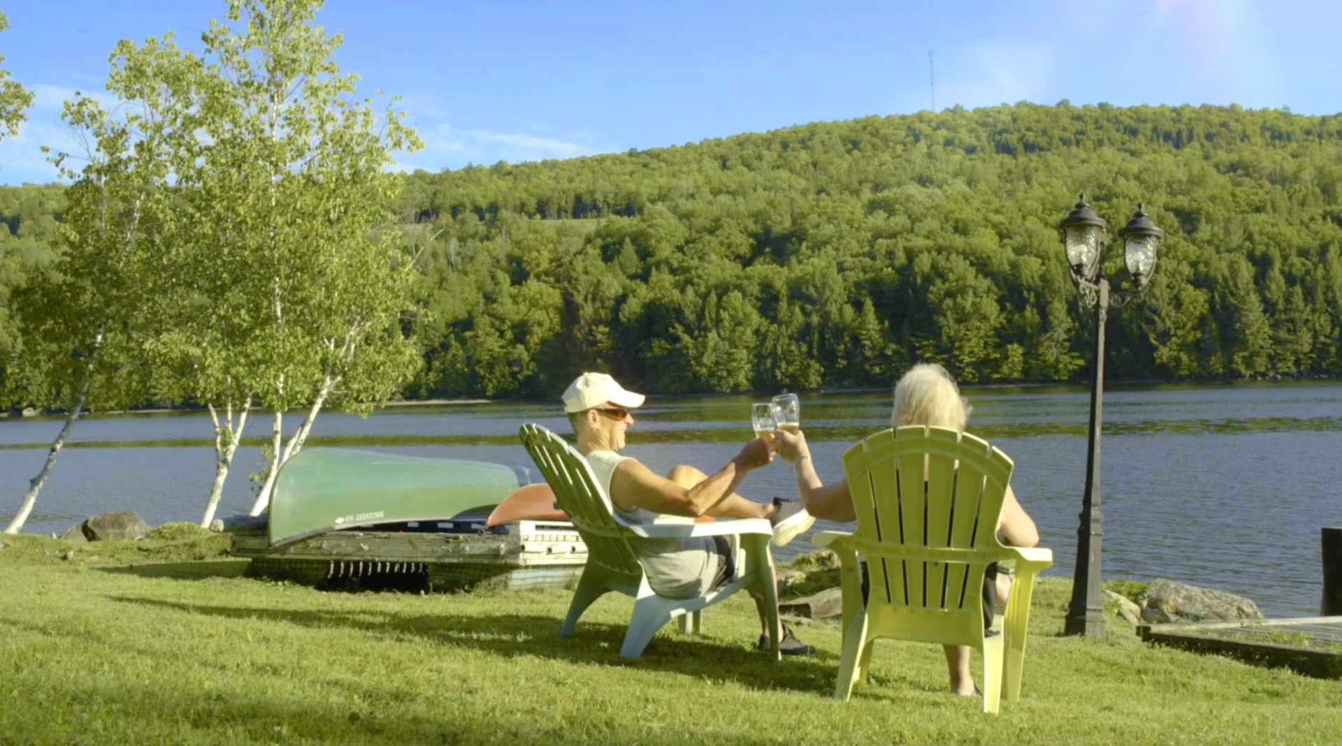 Two people clink glasses in Adirondack chairs on a lawn by a lake, with a wooded mountain in the background.