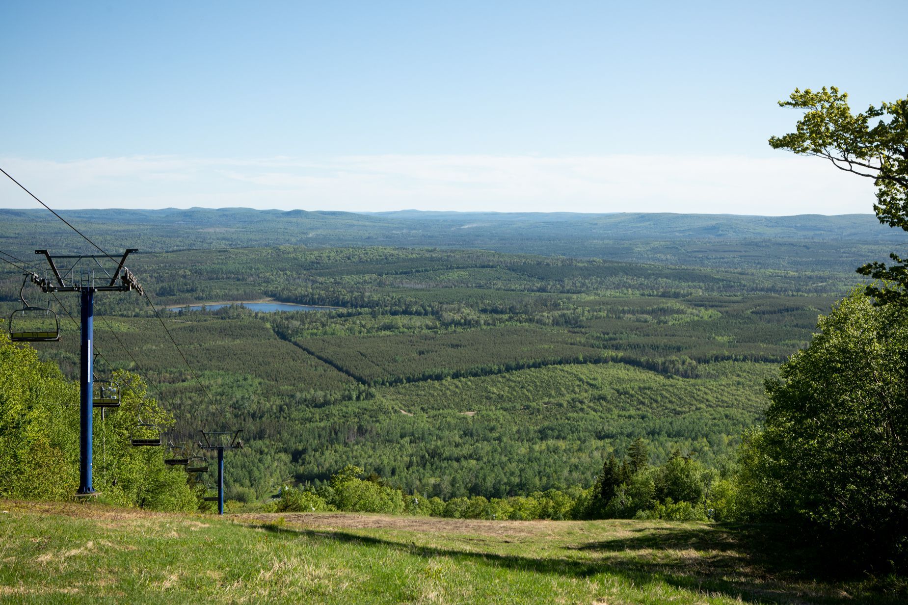 View from a mountaintop, overlooking a vast green forest and a small lake, with a blue ski lift.