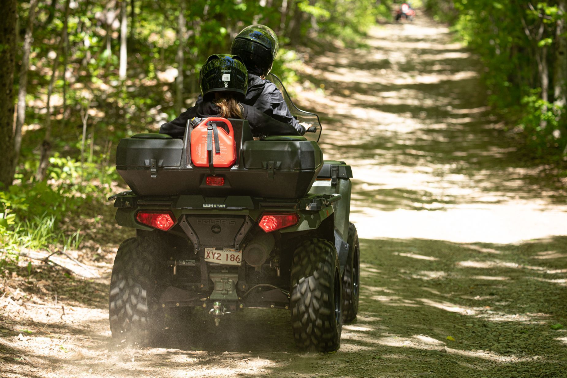 ATV with two riders on a dirt path in a wooded area. Black helmets, storage boxes.