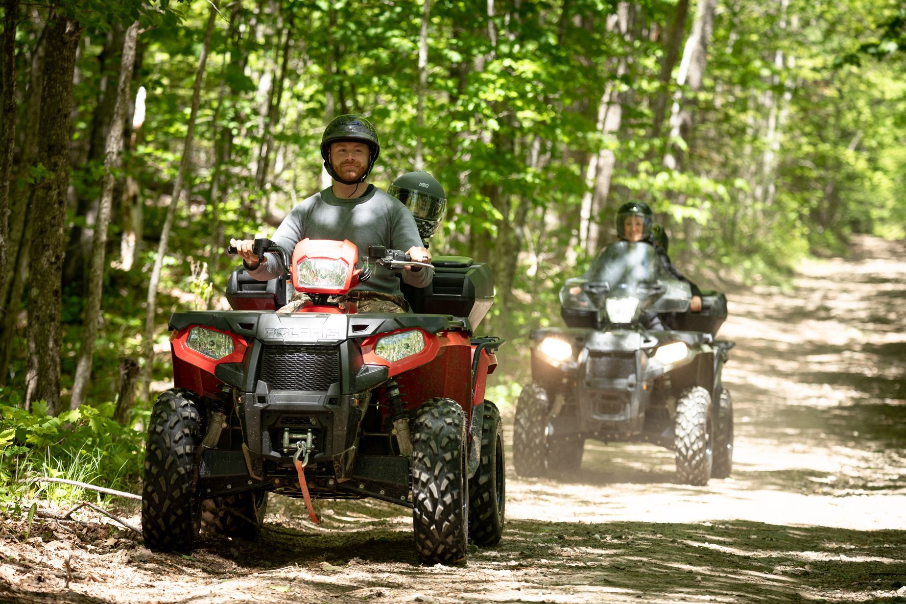 Two people riding ATVs on a dirt path through a forest. The lead rider smiles, the ATV is red.