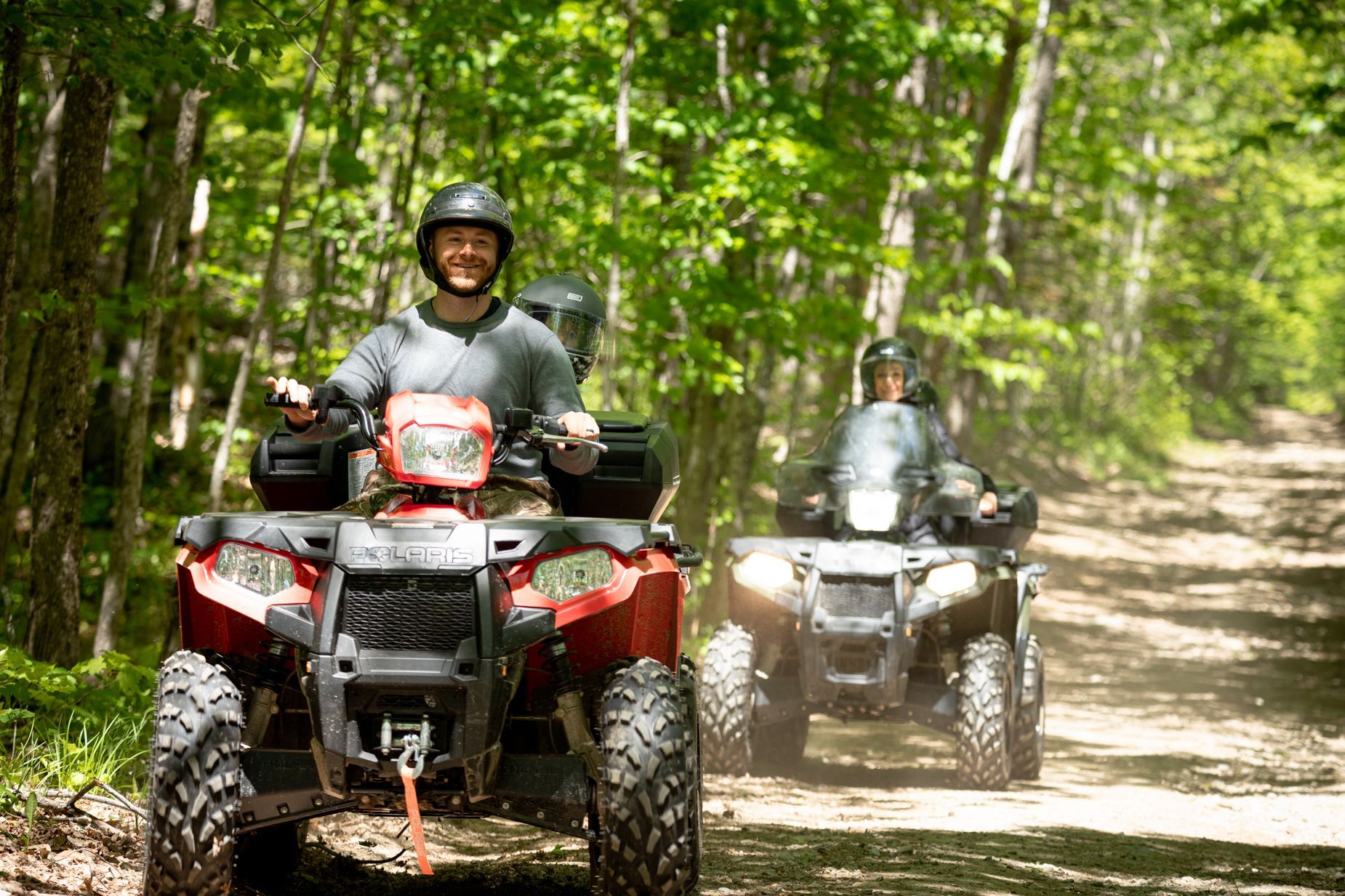 Two people ride ATVs on a dirt path through a sunny forest. One ATV is red.