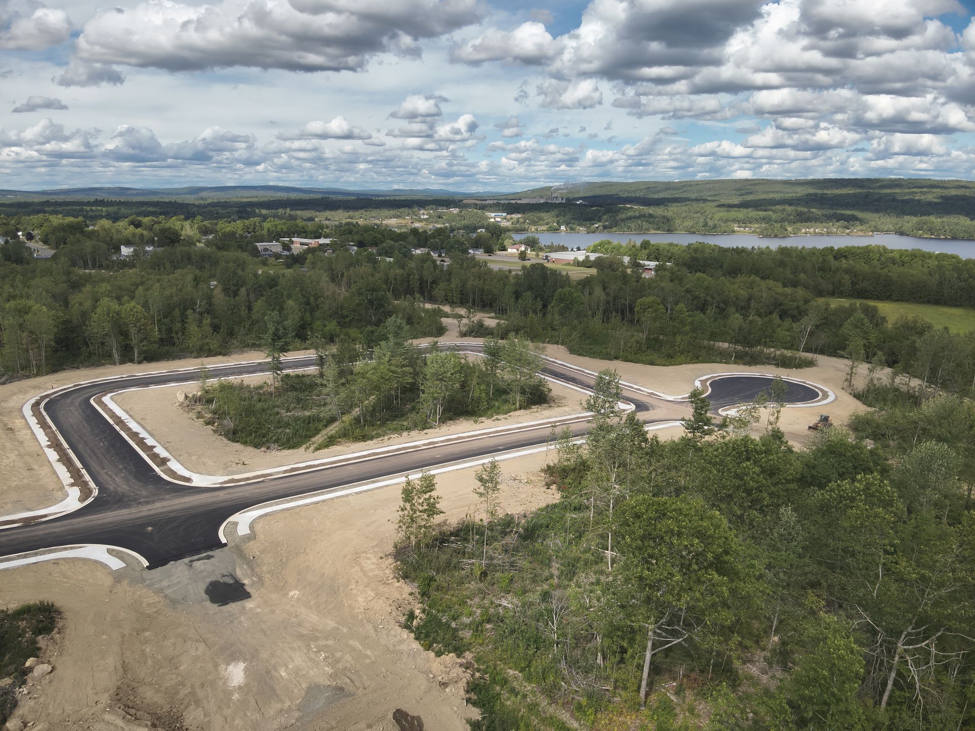 Aerial view of a new road and parking area under construction beside a wooded landscape and cloudy sky