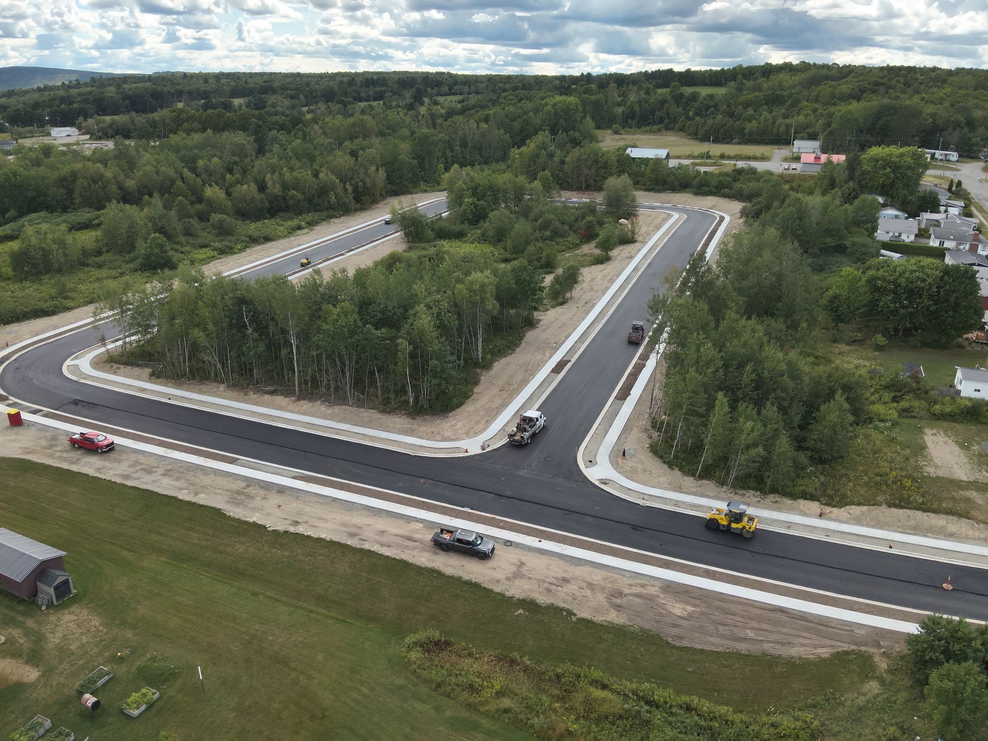 Aerial view of a new road intersection with paved lanes, trees, and a few vehicles under cloudy sky