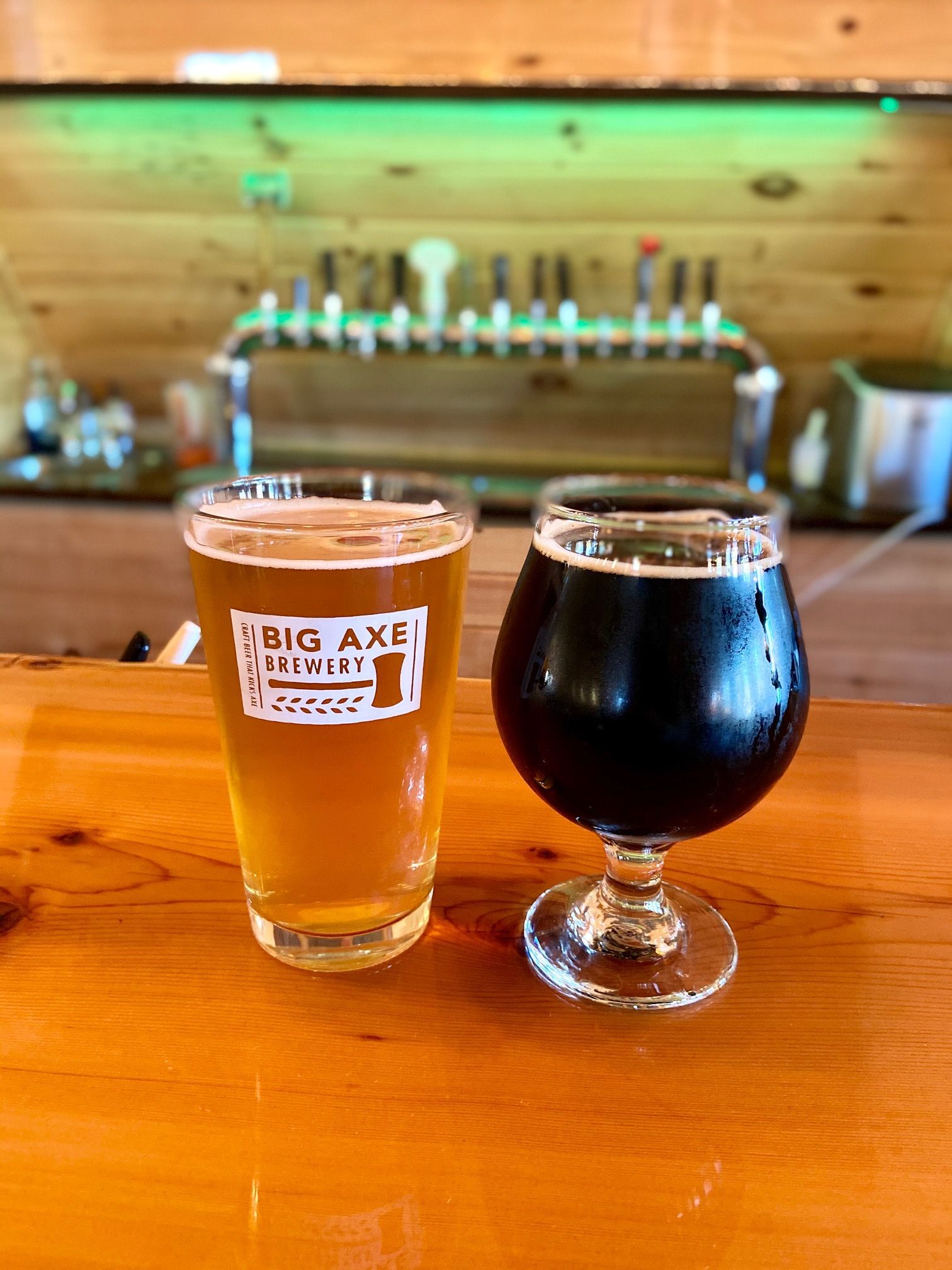 Two beers on a wooden bar at Big Axe Brewery, one light, one dark. Bar taps in background.
