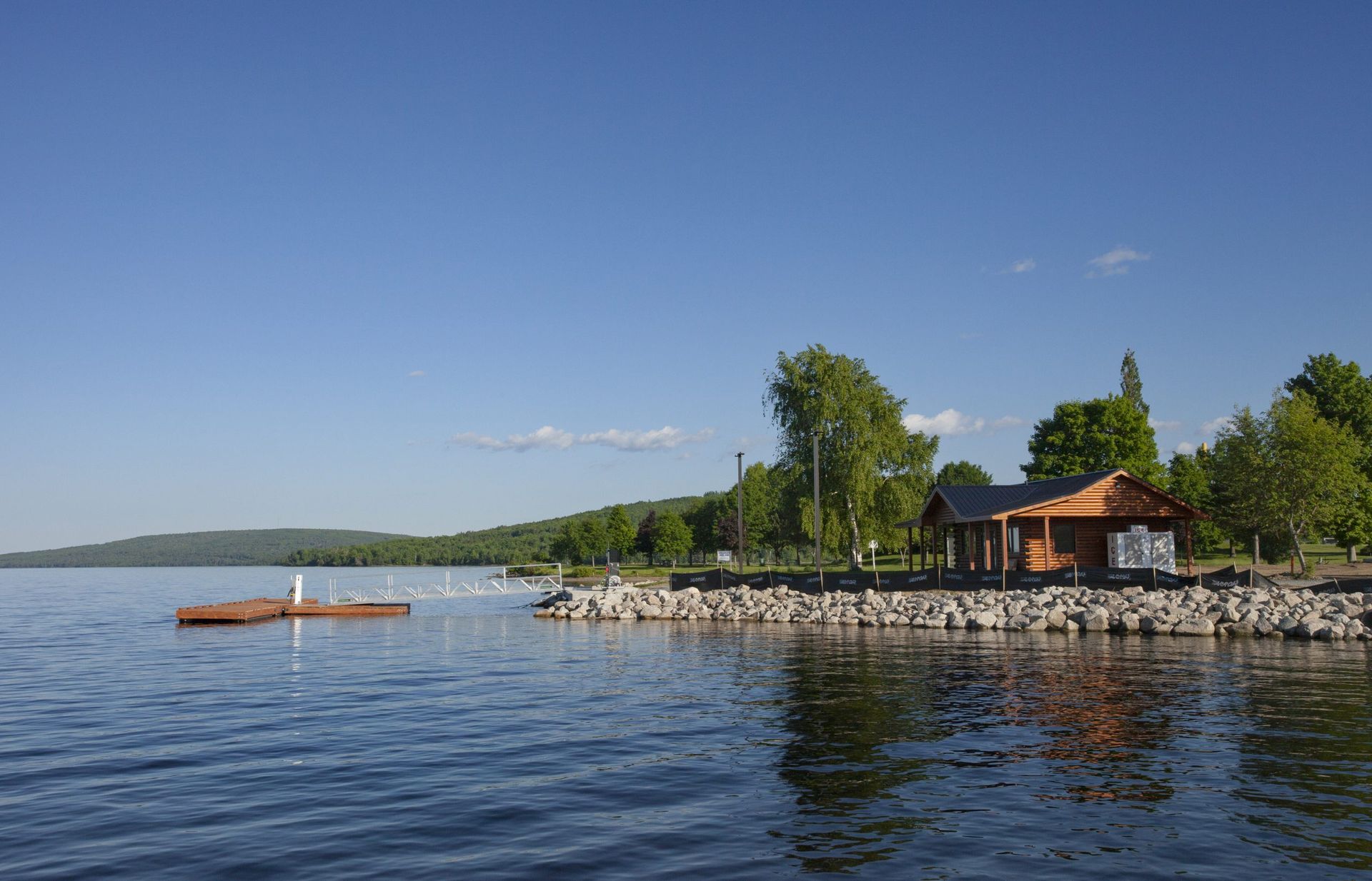 Lakeside scene with small wooden building, dock, trees, and distant hills under a blue sky.