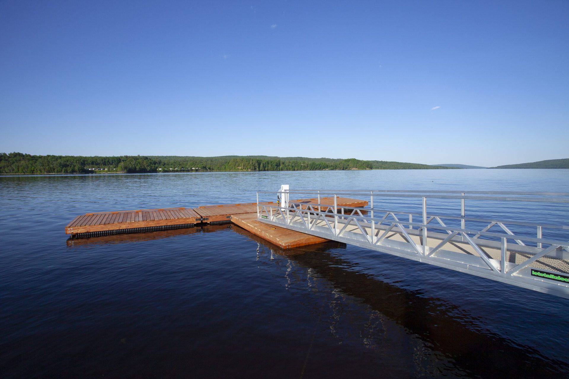 Dock extending into a lake, with a ramp. The dock is brown, the ramp is silver, and the sky is blue.