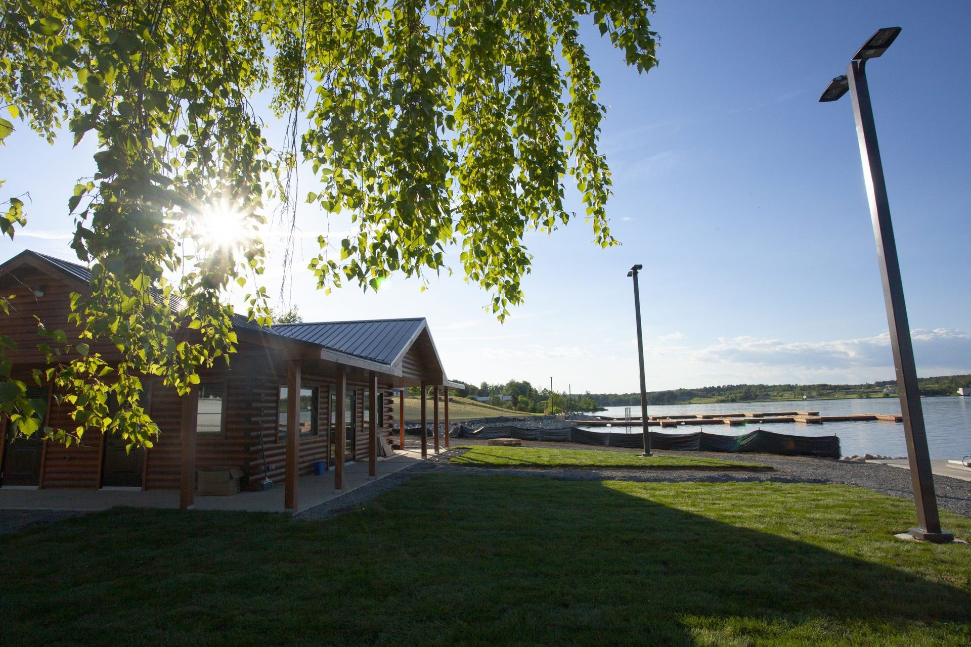 Lakeside log cabin with a porch, green lawn, sunlit trees, and a distant pier under a blue sky.