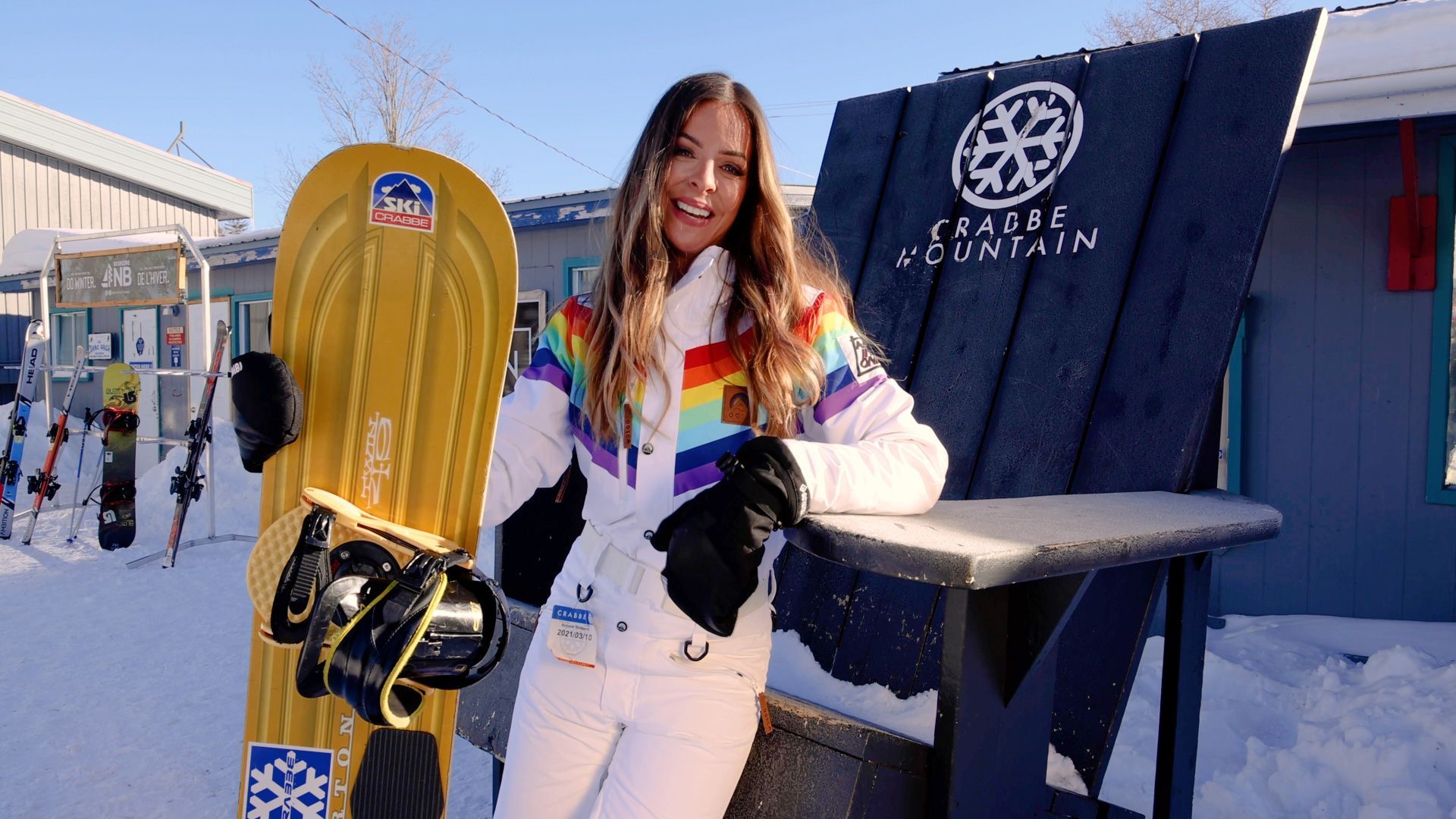 Woman in rainbow ski jacket smiles next to a yellow snowboard outside a building with the 