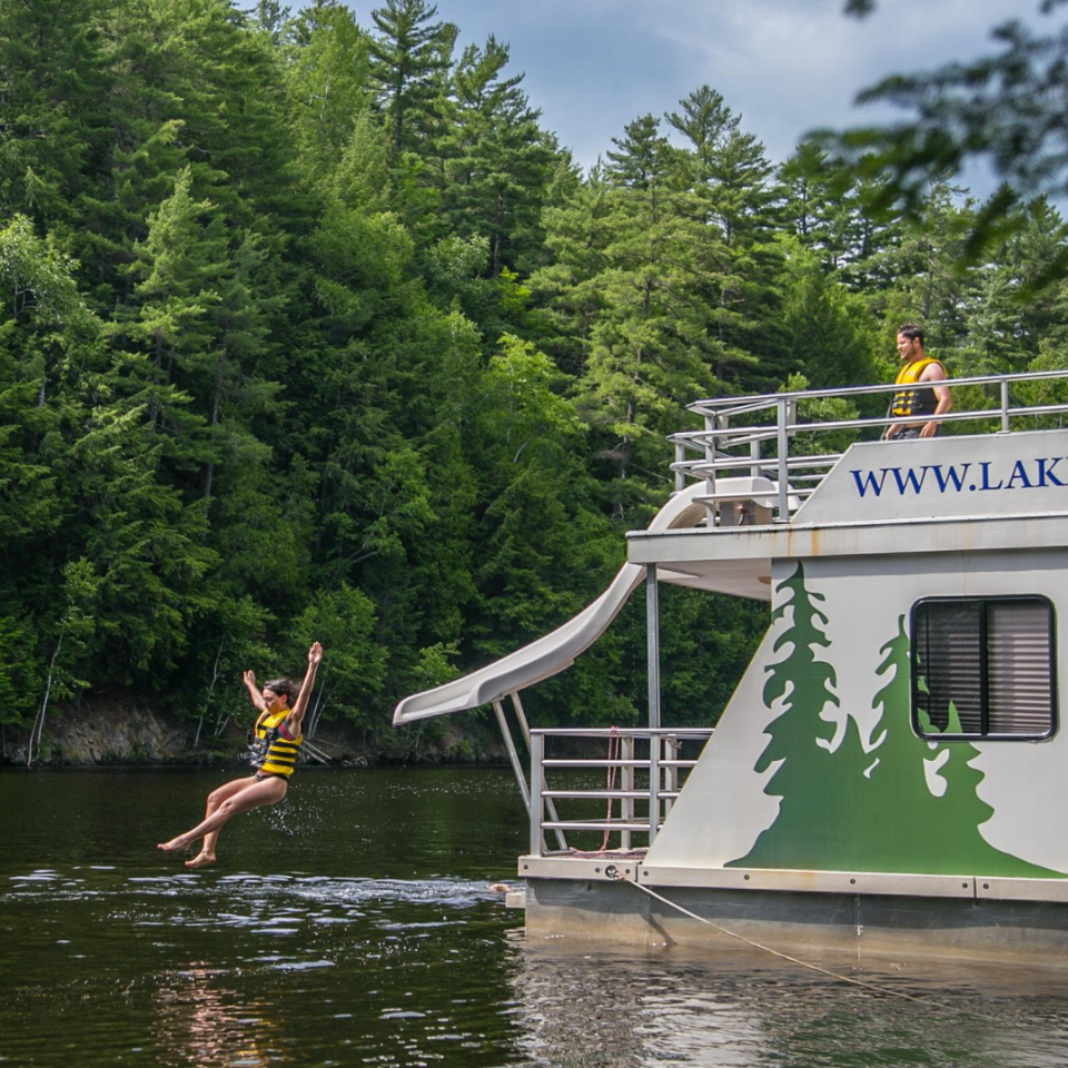 Person jumping from a houseboat into a lake, wearing a life vest. Another person stands on the upper deck.