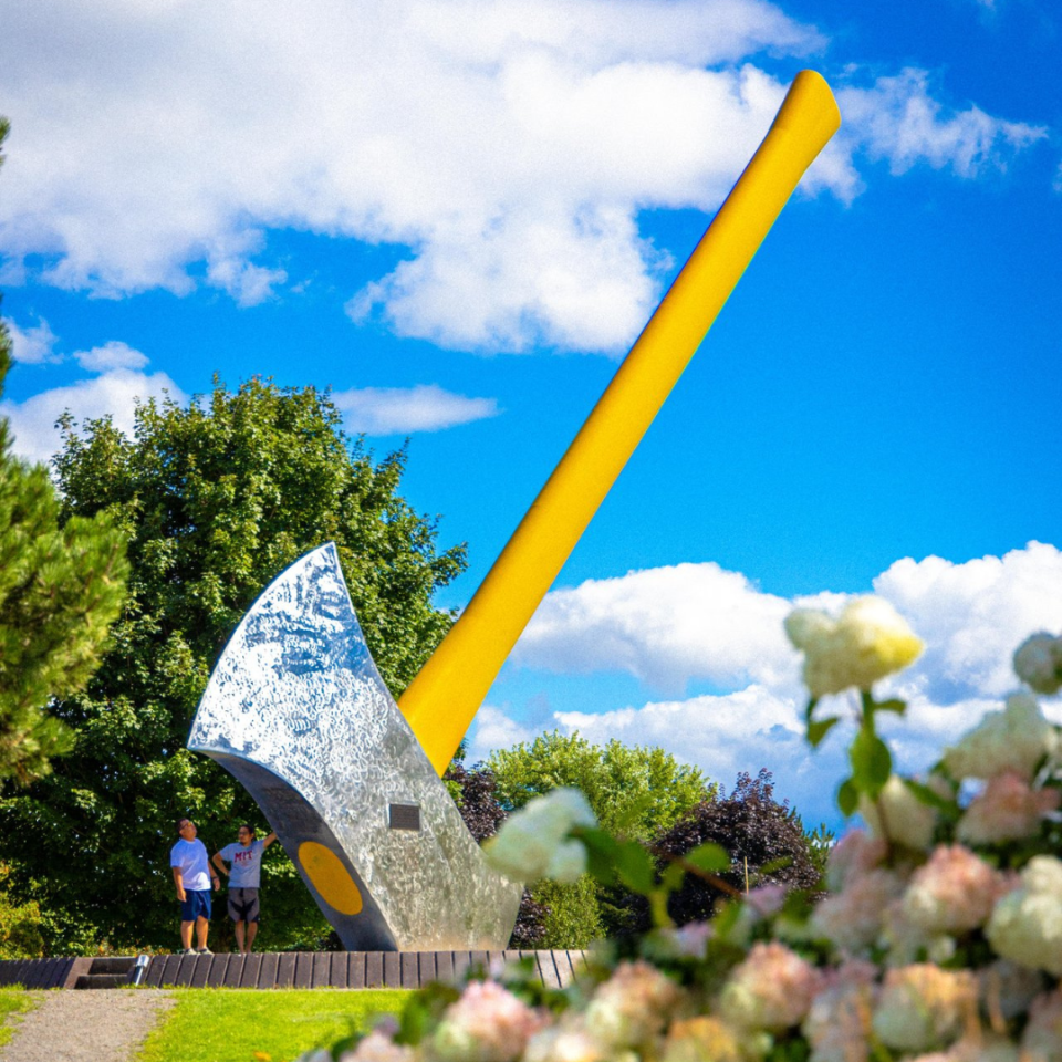 Large yellow-handled axe sculpture with silver blade in a park with two people, blue sky, and flowers.