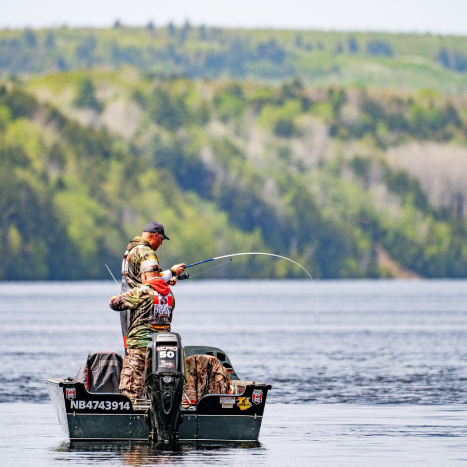 Two people fishing from a boat on a lake with treed hills in the background.
