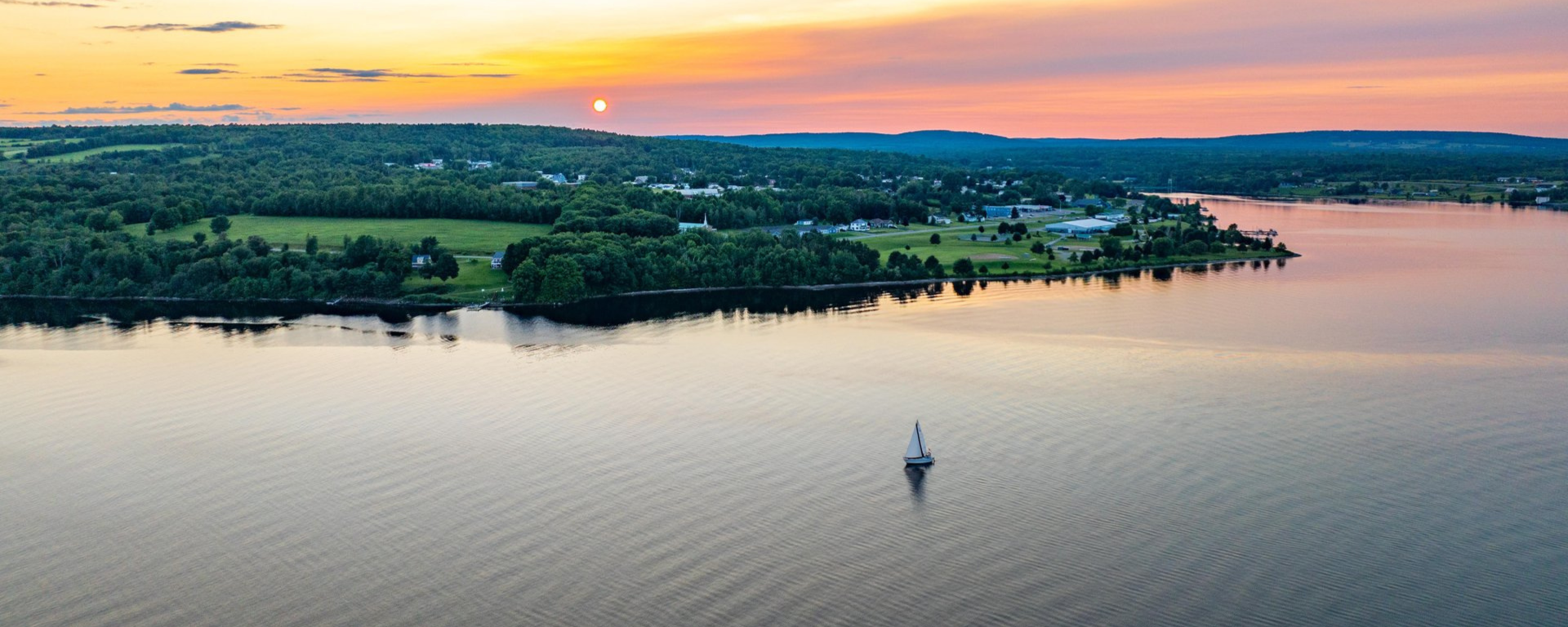 Sailboat on calm water with a forest shoreline and colorful sunset sky.