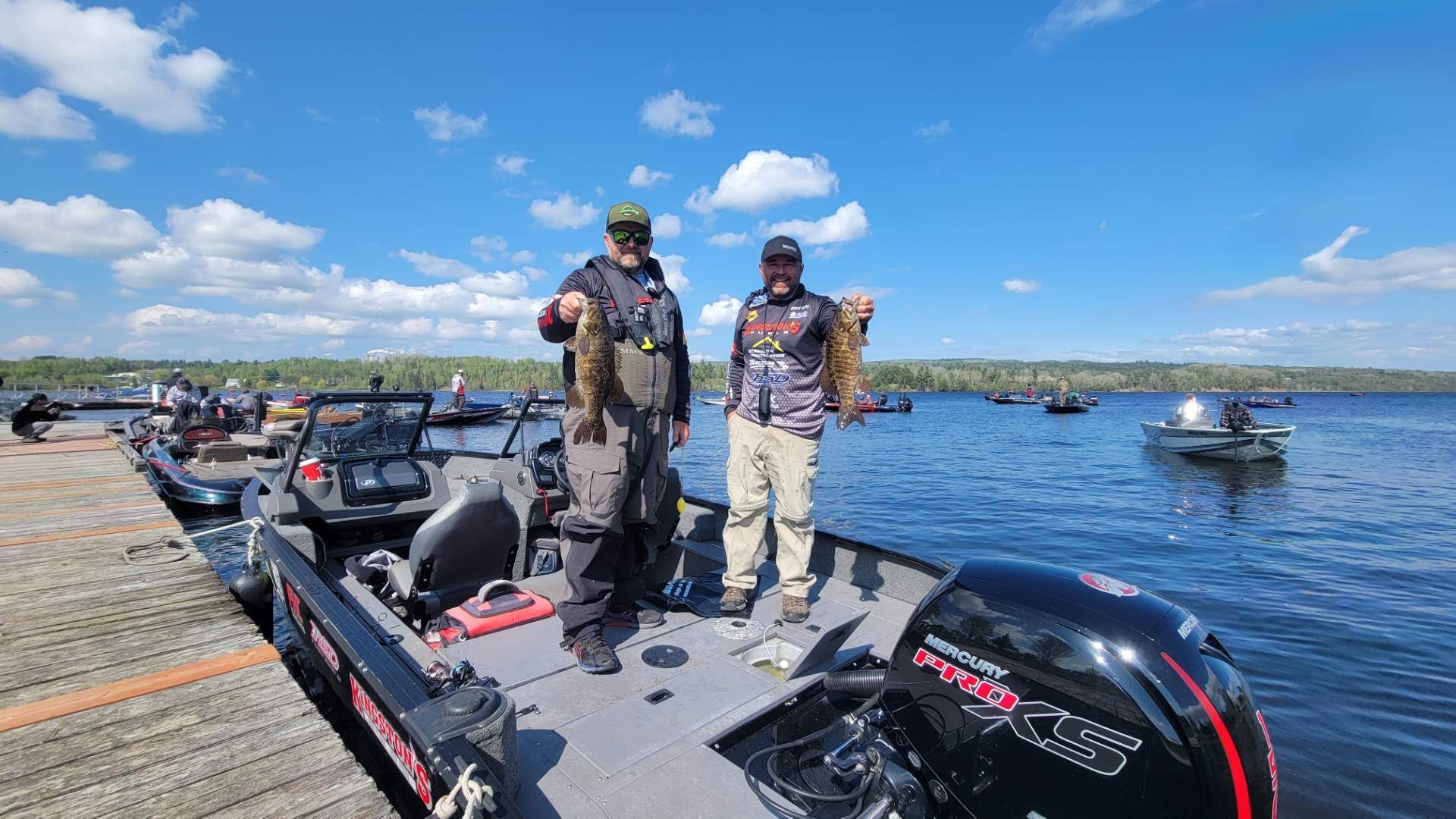 Two anglers on a boat holding fish, blue sky, other boats in the distance, sunny day.