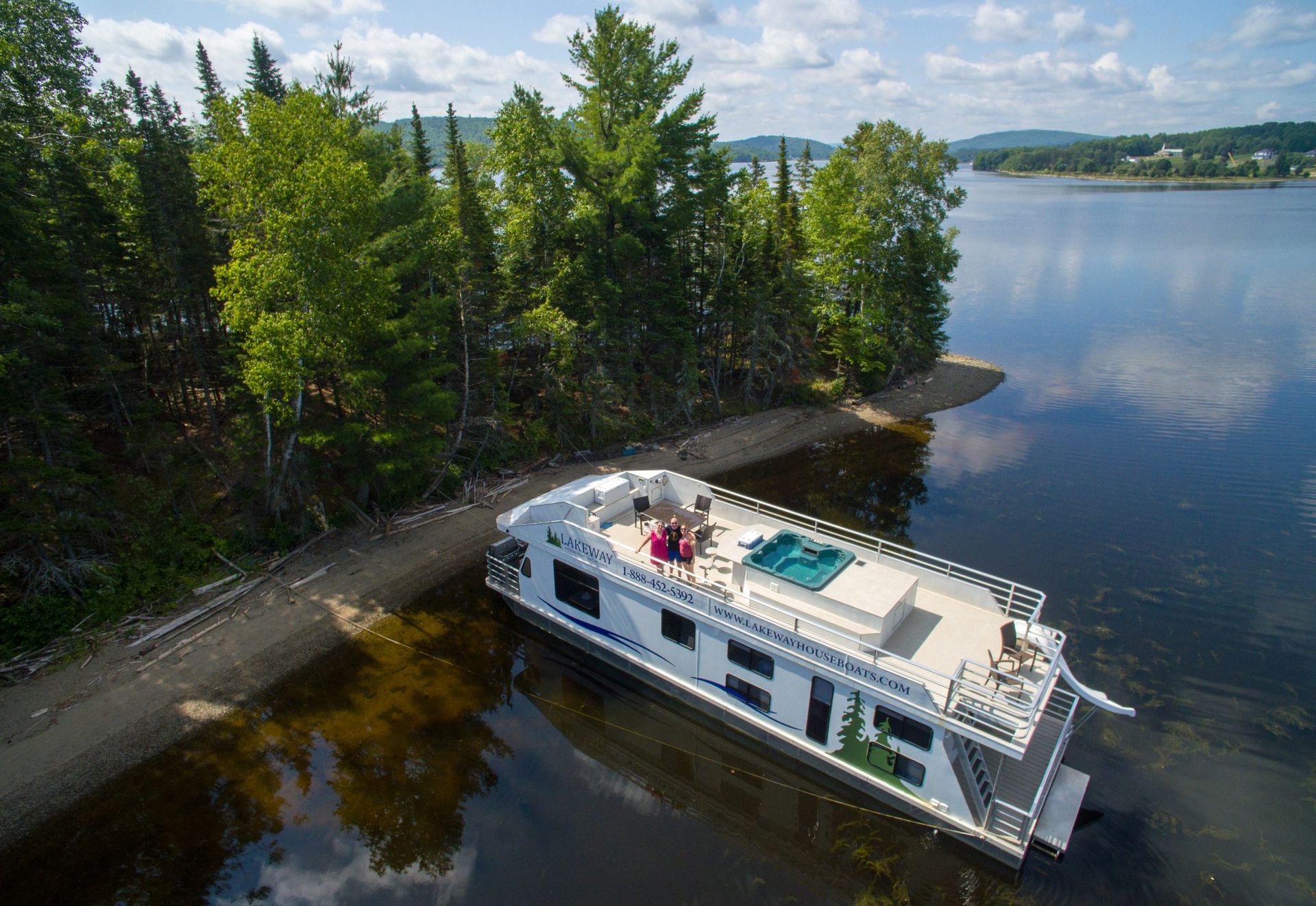 Houseboat on a lake shore. White structure with a hot tub, trees on bank, blue water.