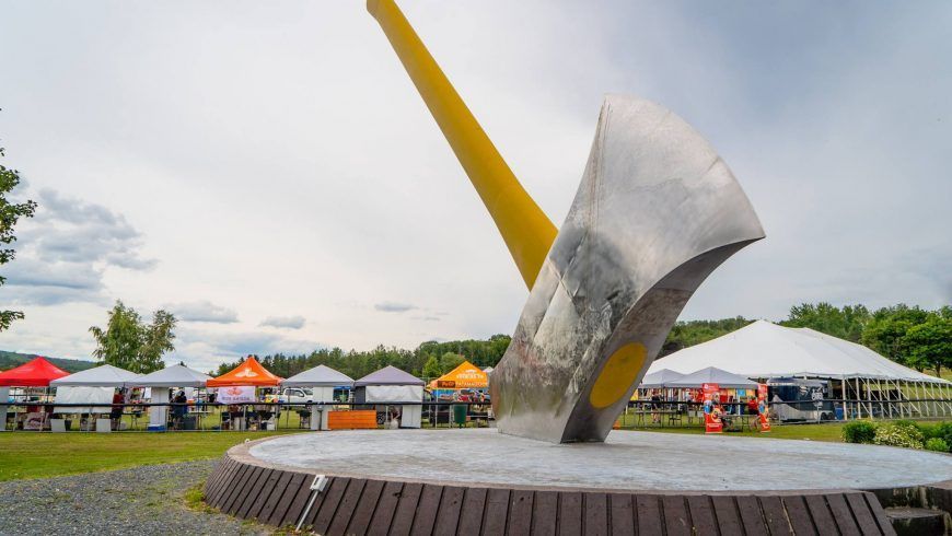 Giant silver axe sculpture with a yellow handle, outdoor event with tents in background.
