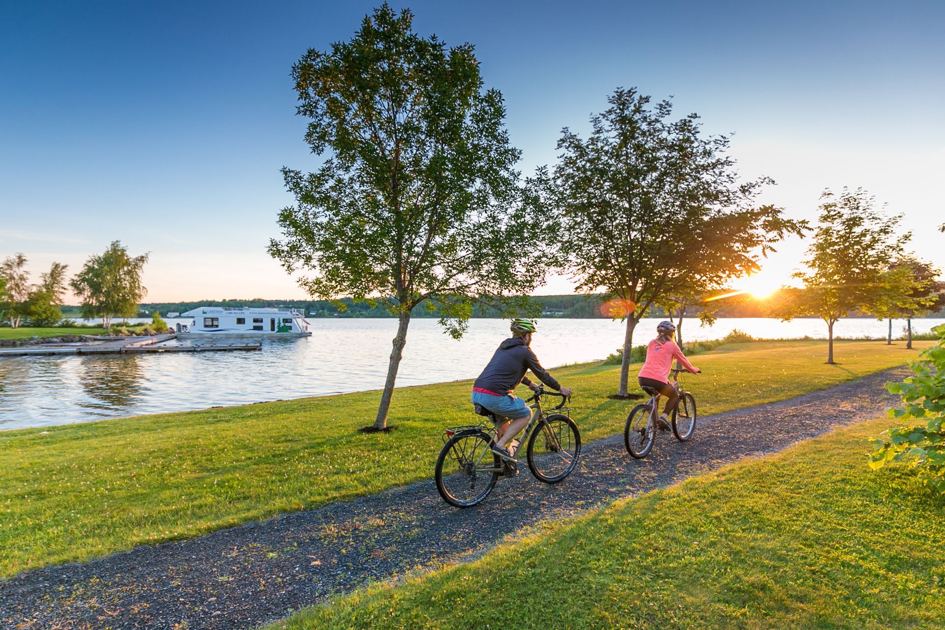 Two cyclists on a path beside a lake at sunset. Green grass, trees, and a house boat visible.