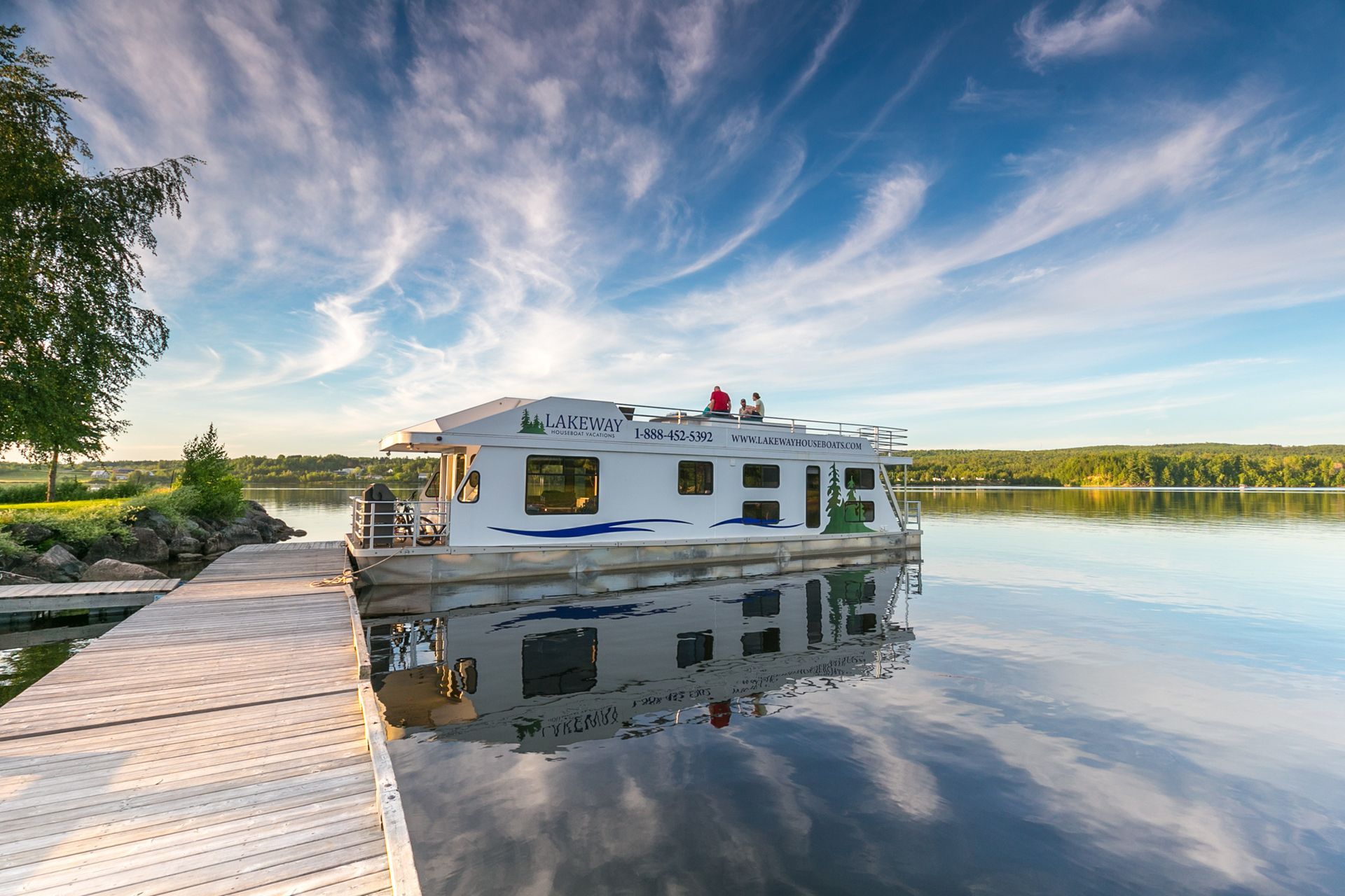 Houseboat on calm lake, docked at wooden pier; blue sky with clouds reflected in water.