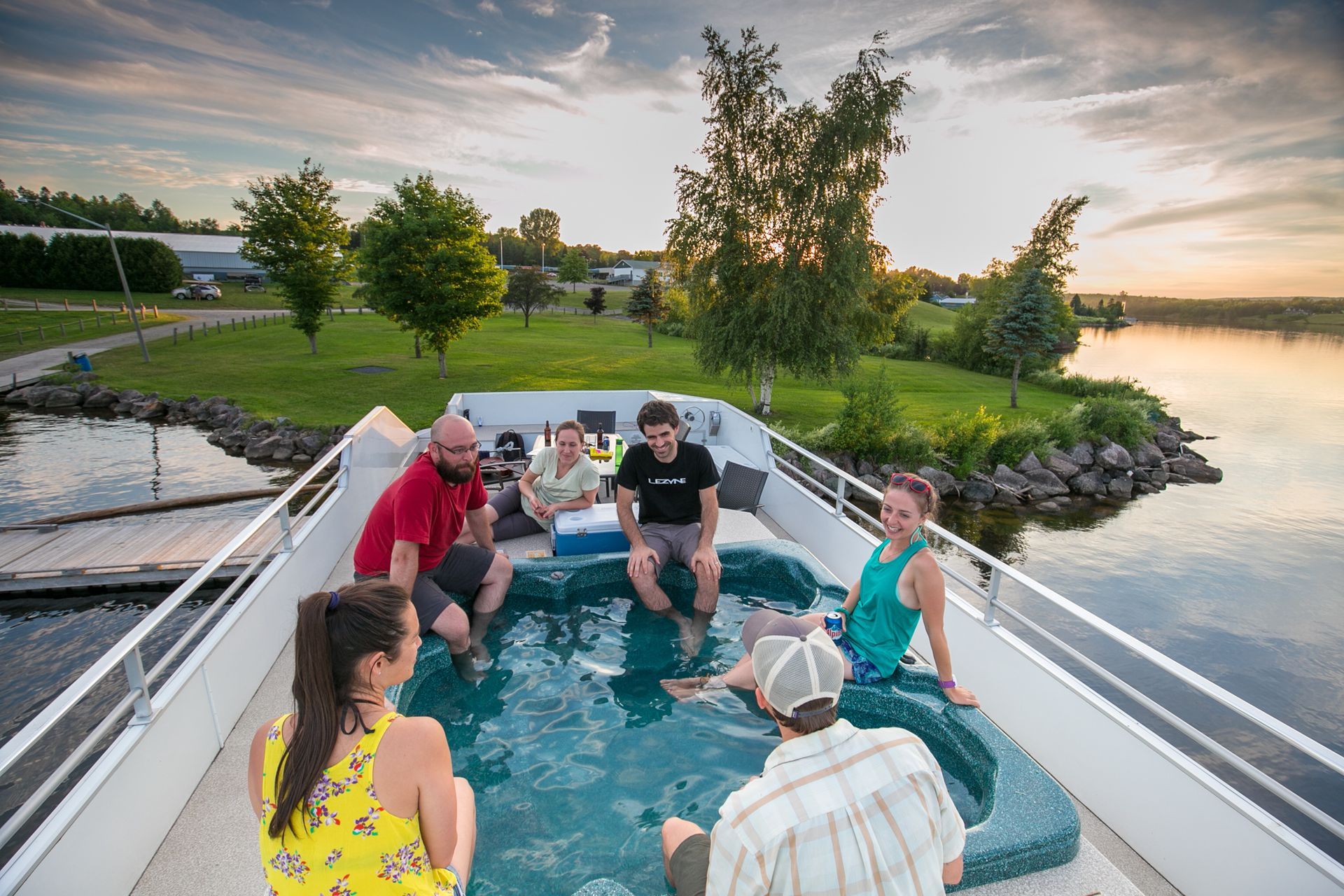 Group relaxing in a hot tub on a boat, overlooking a lake at sunset.