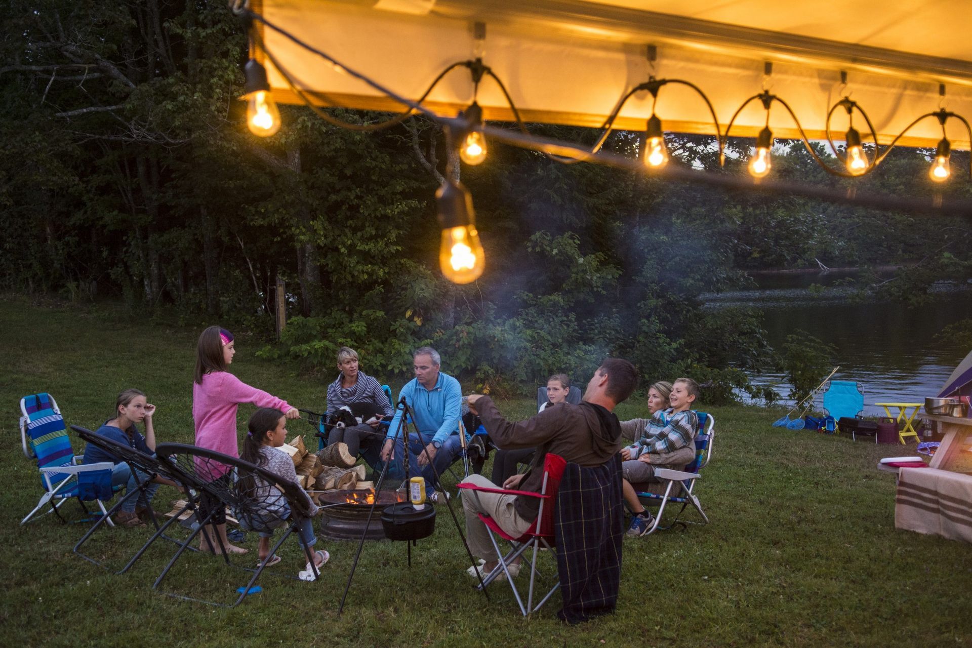 People gathered around a campfire at dusk, illuminated by string lights.