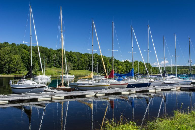 Sailboats docked at a marina, with tall masts reflecting in calm water, under a blue sky.