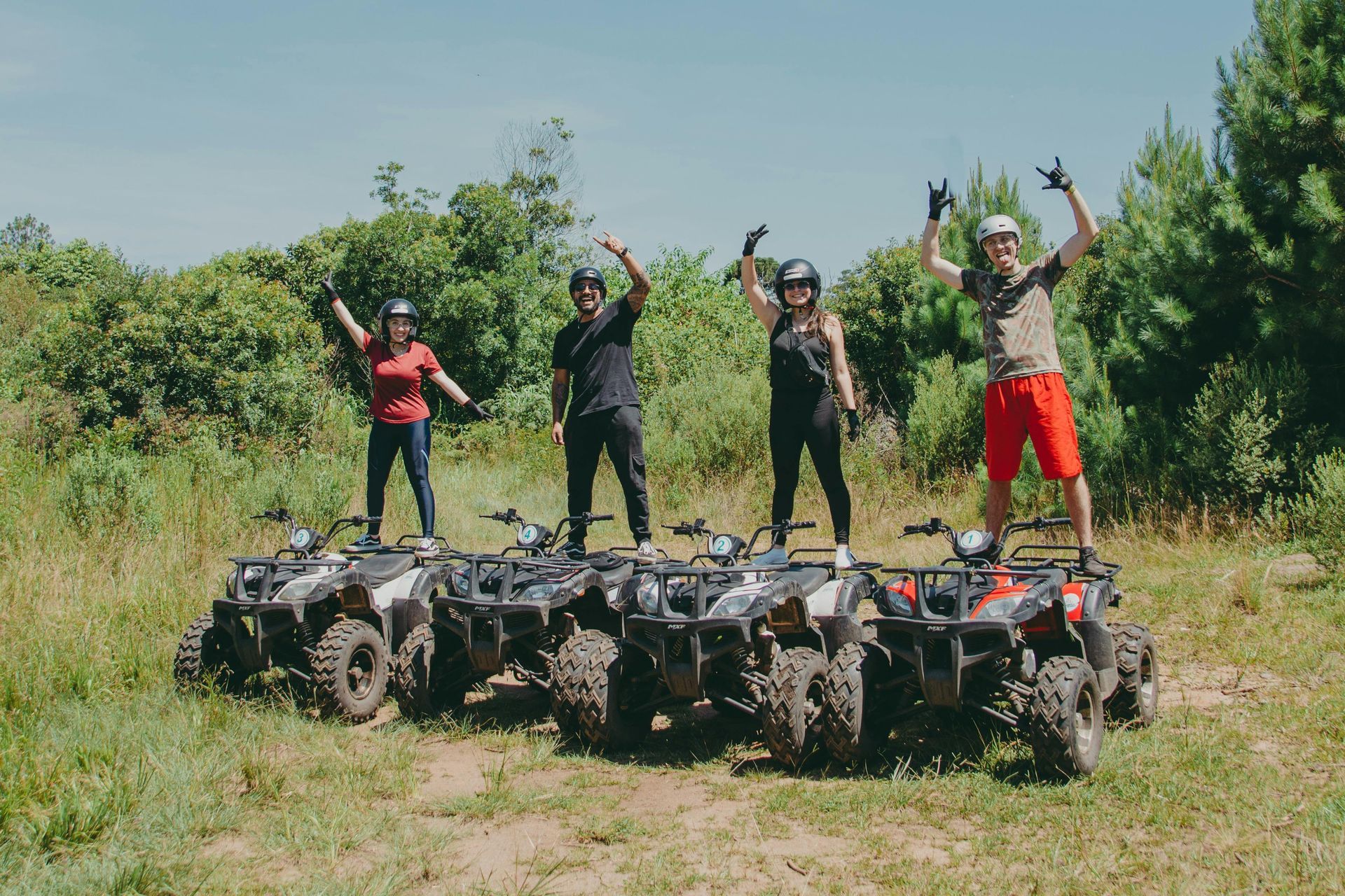 Four people standing on ATVs in a grassy area, arms raised in a celebratory gesture.