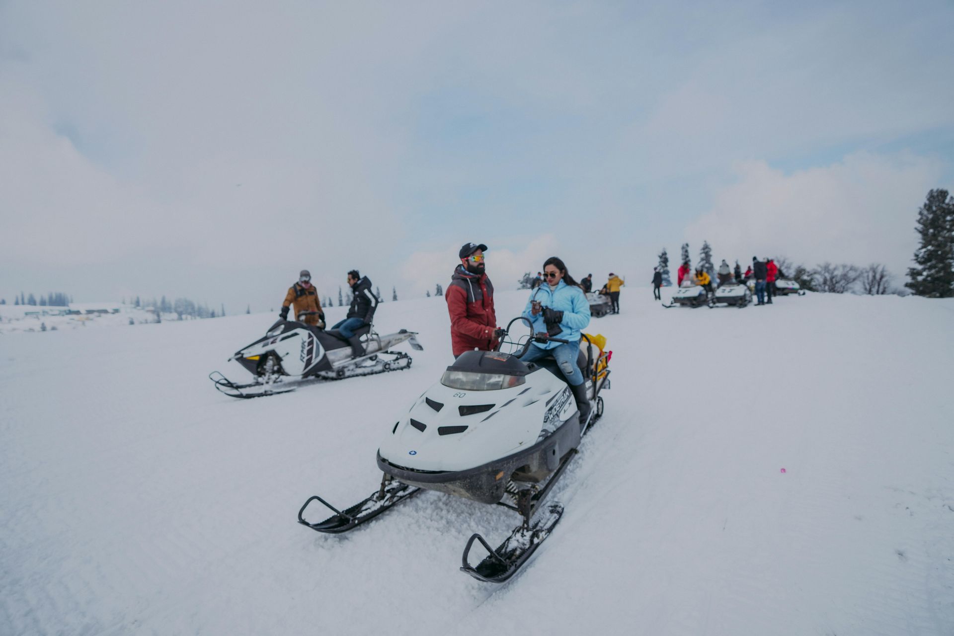 People riding snowmobiles on a snowy plain. White snow covers the ground, light blue sky above.