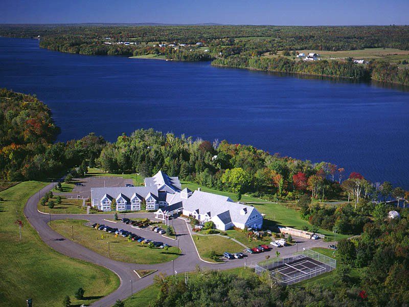 Building near a lake, surrounded by trees; paved driveway and parking. Blue water and sky.