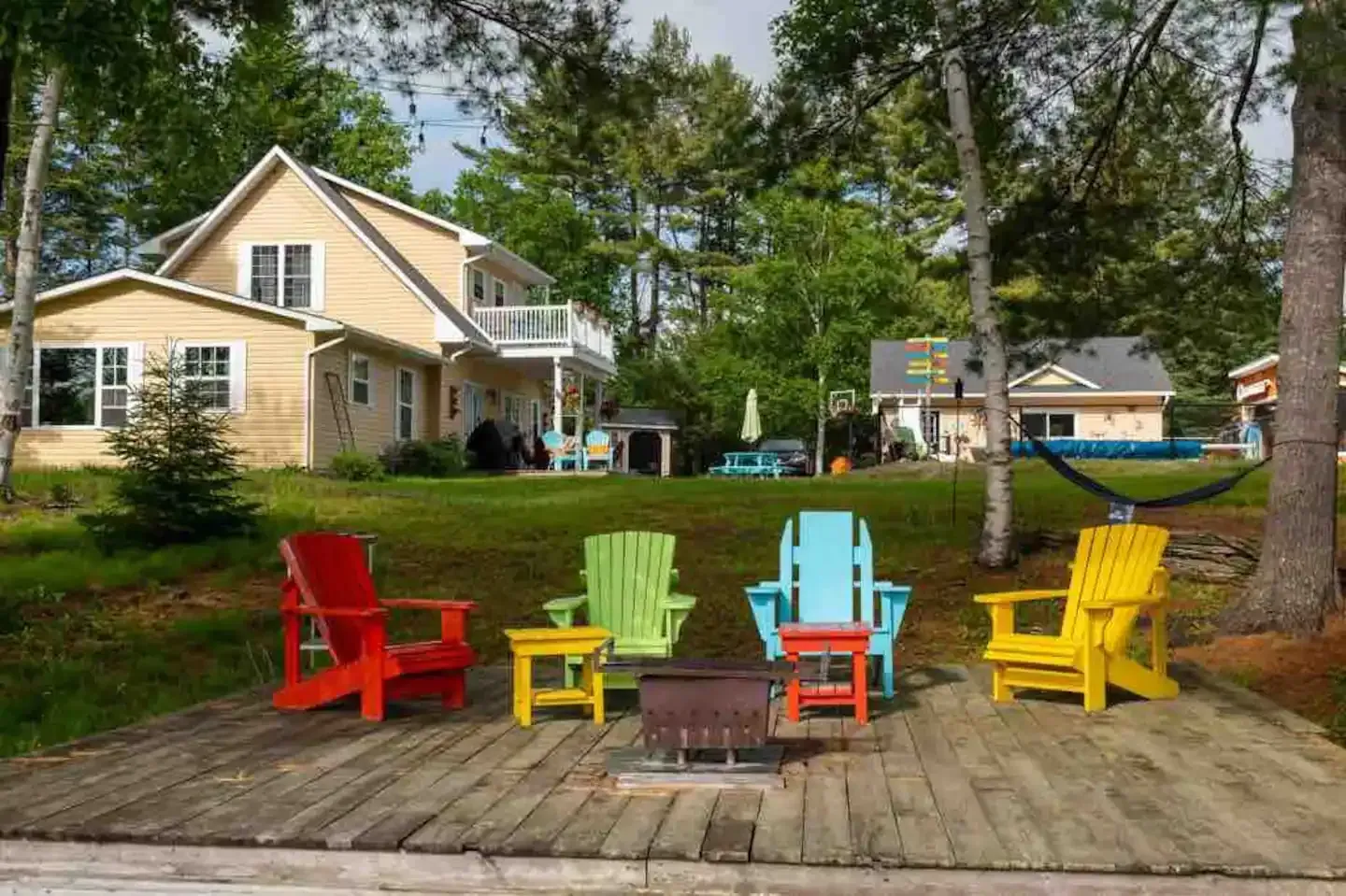 Colorful Adirondack chairs surround a fire pit on a wooden dock in front of a house.