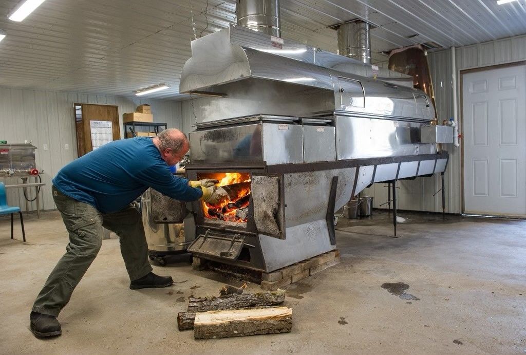 Man adds wood to a fire beneath a stainless steel evaporator, used to make maple syrup. Inside a shed.