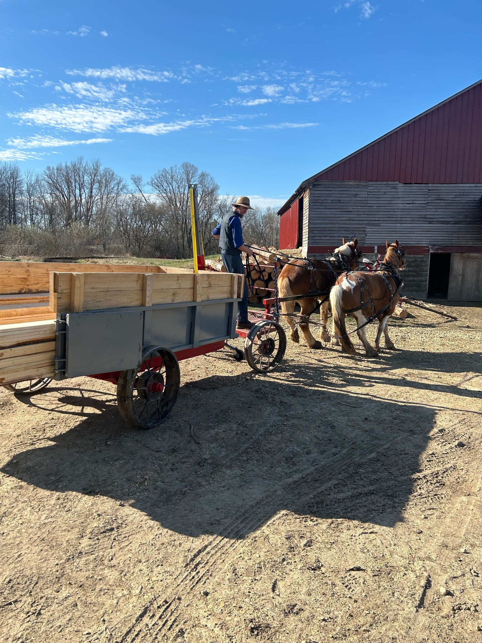 A man is standing next to a horse drawn wagon in front of a barn.