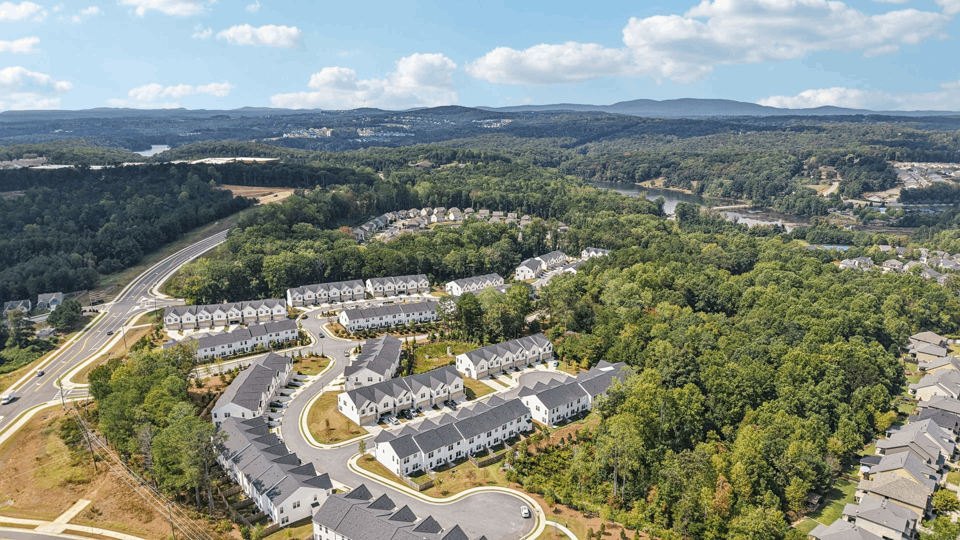 Aerial view of a wooded suburban neighborhood with winding roads and rows of houses under a blue sky.