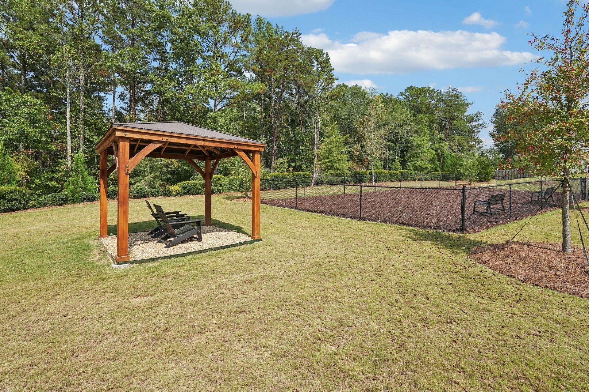 Wooden pergola with a bench on a grassy yard beside a chain-link fence and trees under a blue sky