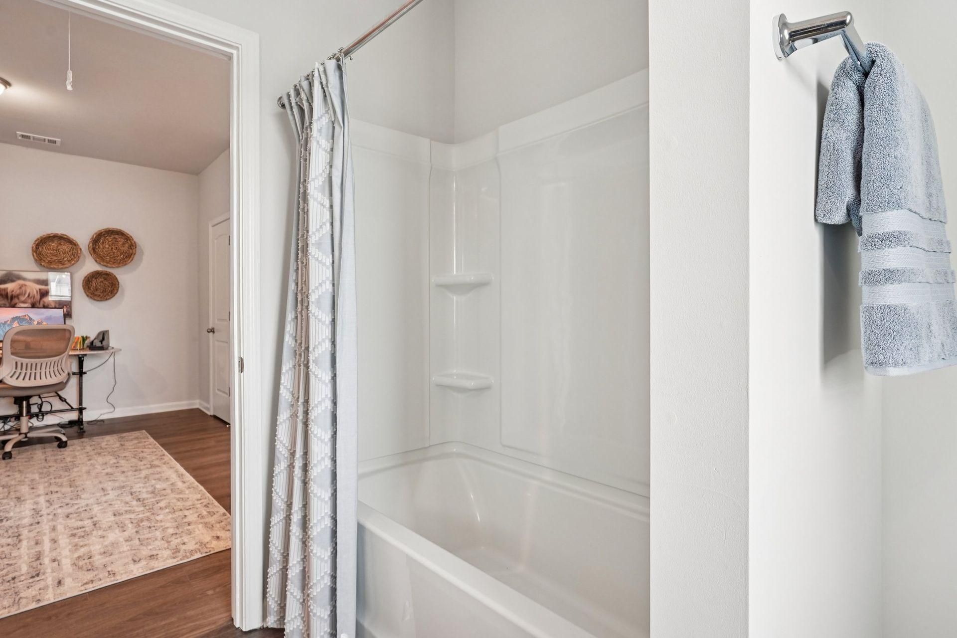 Bathroom shower with clear curtain and blue towel on hook, leading into a room with rug and table