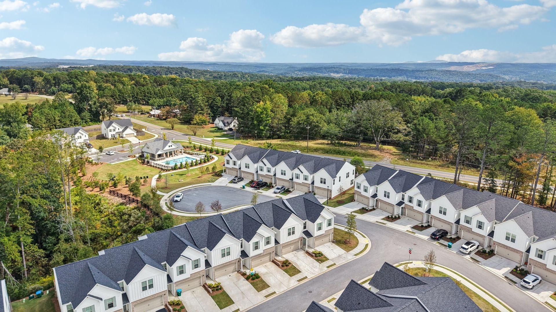 Aerial view of a suburban apartment complex with gray roofs, trees, and distant city skyline under blue sky