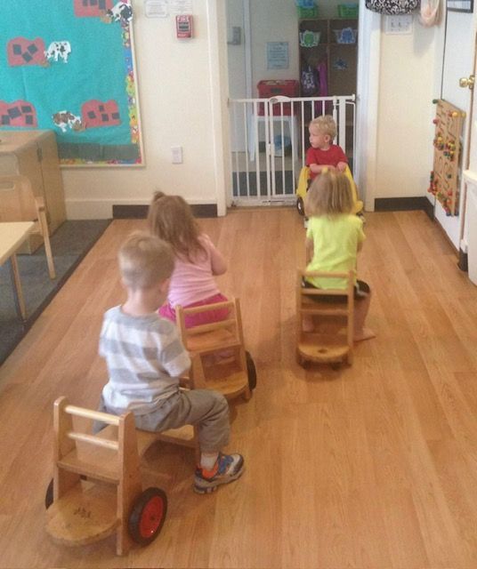 Children riding wooden scooters in a daycare room toward a seated child at a gate.