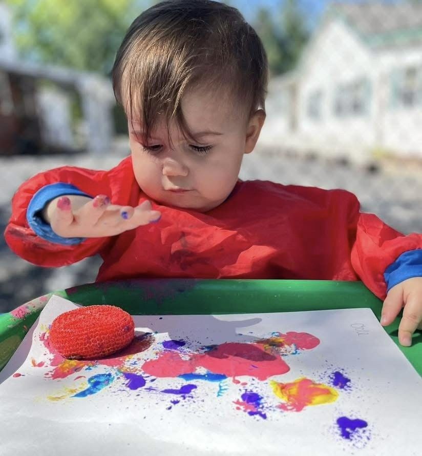 Child in a red shirt looking at paint on their hand while making colorful art at a table.