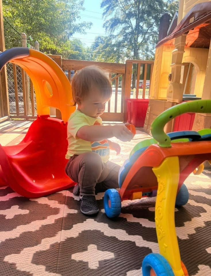 Child sitting on a colorful playset outdoors, with a red slide and patterned play mat.