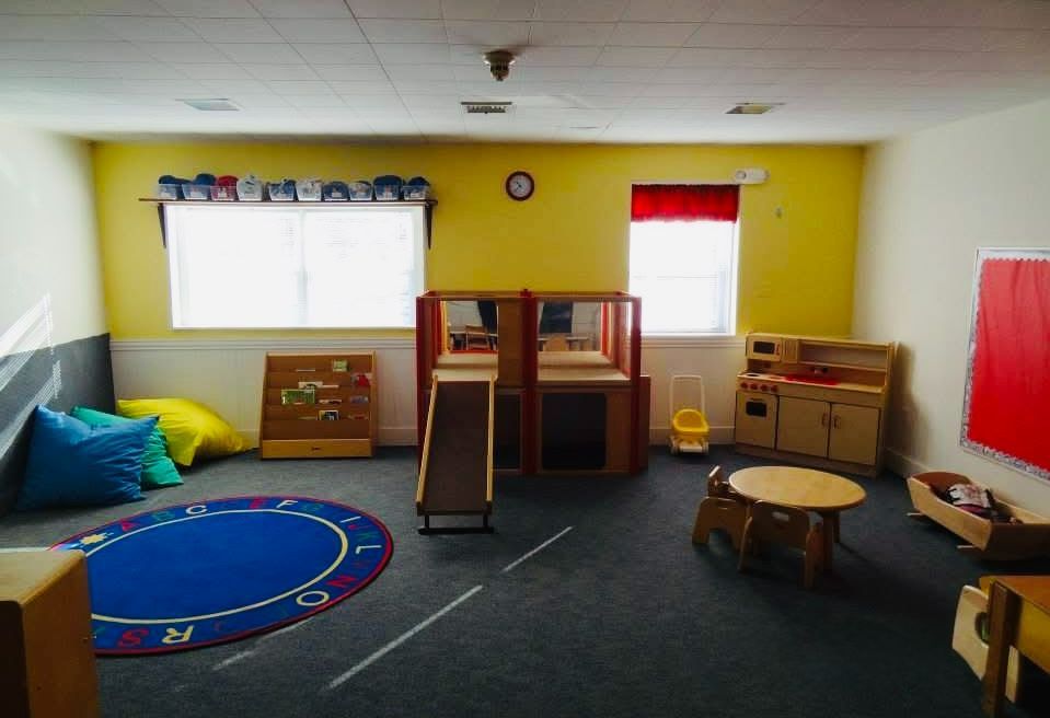 Bright preschool classroom with yellow walls, play rugs, toy shelves, and small tables under windows
