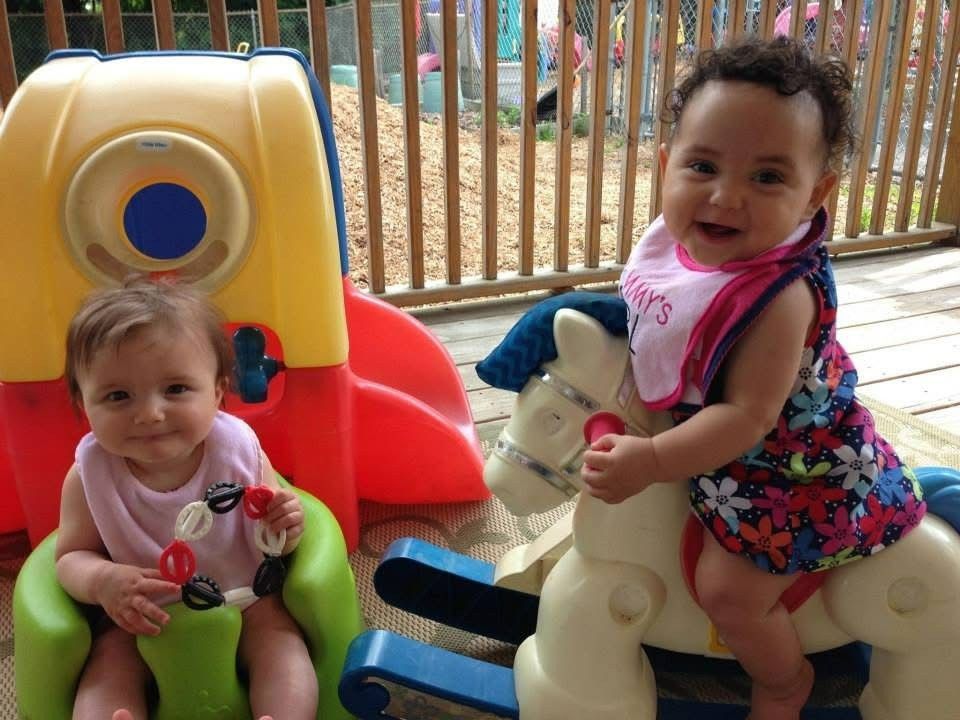 Two toddlers on plastic ride-on toys at a playground, smiling in colorful clothes.