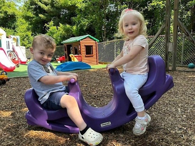 Two children riding a purple rocking toy at a playground, with a playhouse and slides in the background