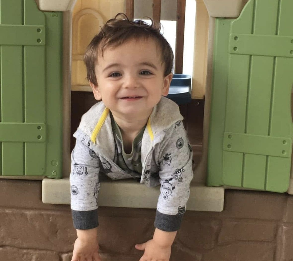 Smiling toddler leaning out of a playhouse window with green shutters