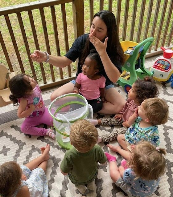 Adult sitting with several small children on a porch, blowing bubbles into a large bubble container.