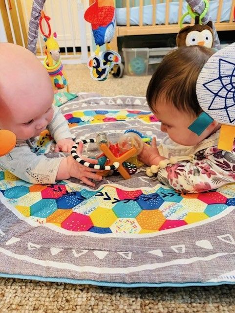 Two babies lying on a colorful play mat, reaching for toys in a nursery