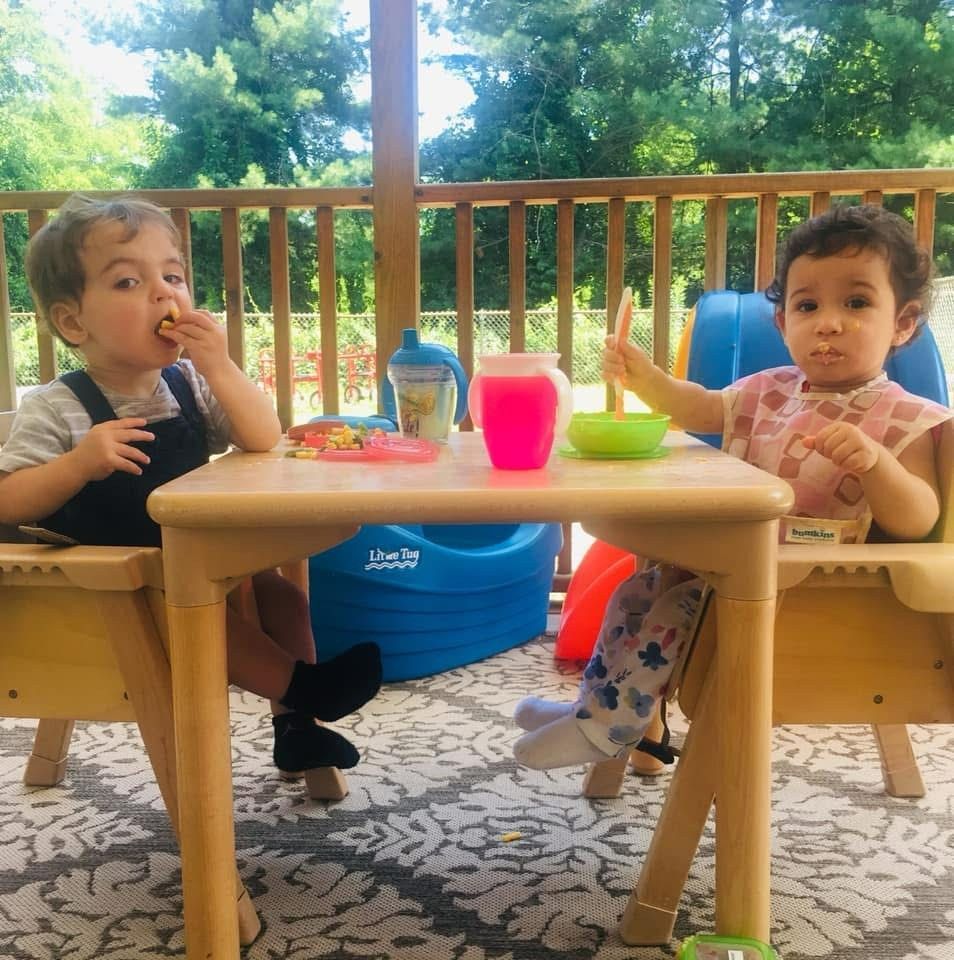 Two children sitting at a table outdoors, eating snacks with colorful cups and bowls.